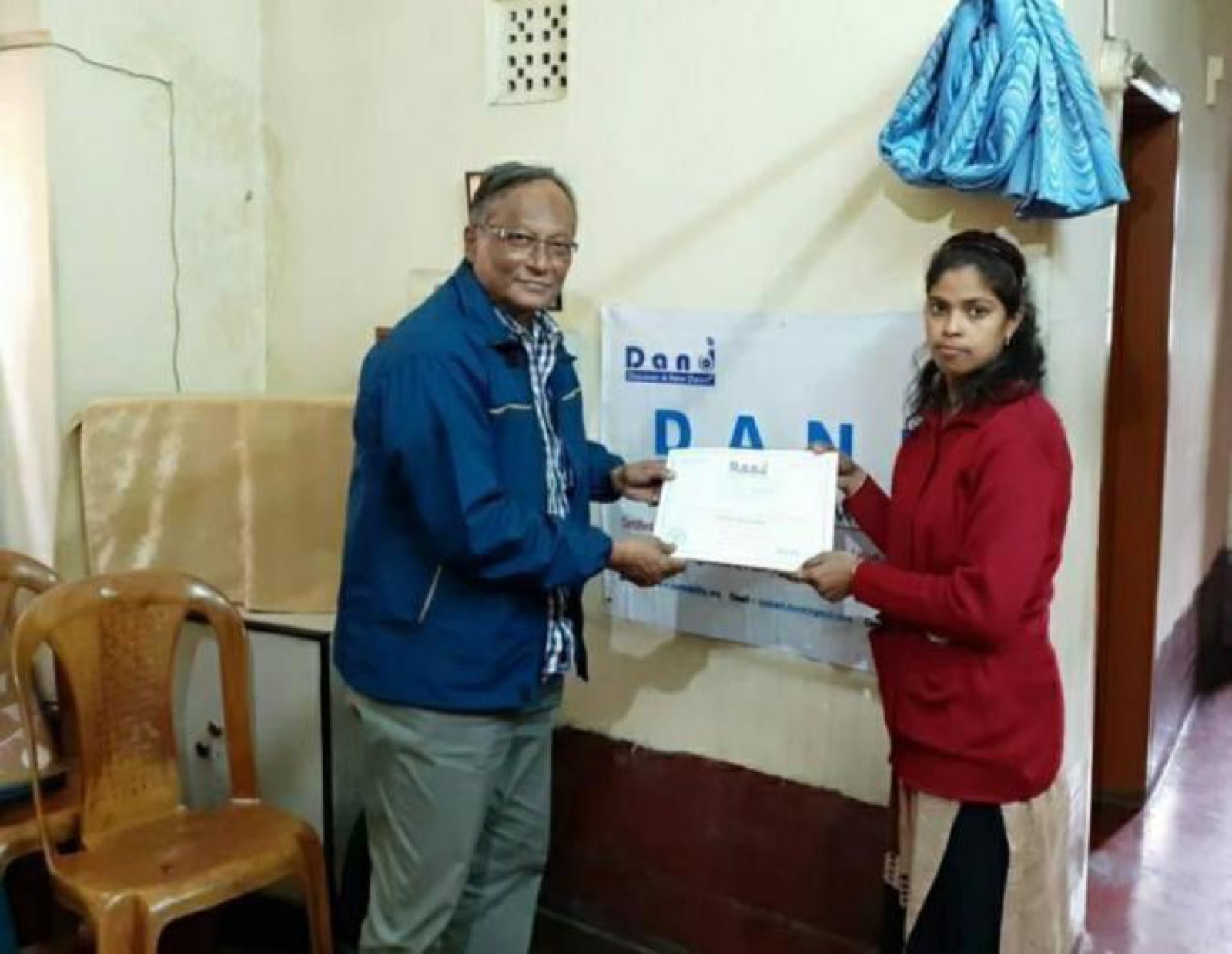 A young woman receives a First Step grant certificate from a DAND supporter in front of the DAND banner.